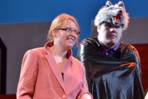 An actor dressed up as a wolf with a wolf headdress on next to a woman wearing a red blazer