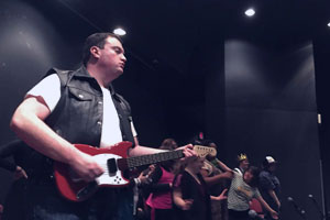 A young man with dark hair wears a white tshirt and leather vest. He is holding a small, red electric guitar. The background is black with beams of light.