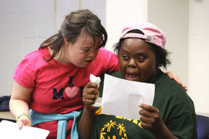Two young women from the waist up in a white room. The woman on the left wears a pink shirt and has dark hair. She leans over to comfort the woman on the right. The woman on the right is holding a piece of paper. Her eyes are wide and she looks very scared.