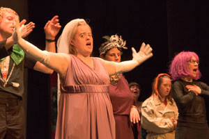 A young woman wearing a pink dress and a white briday veil stretches her arms wide and sings. An eclectic crowd of actors is behind her.