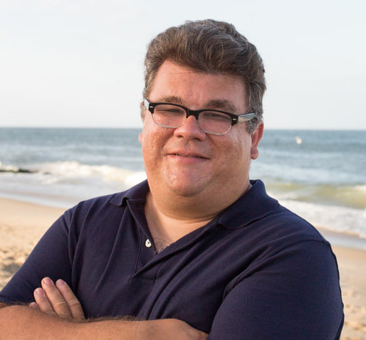 Man in blue polo shirt standing with arms crossed in front of the ocean