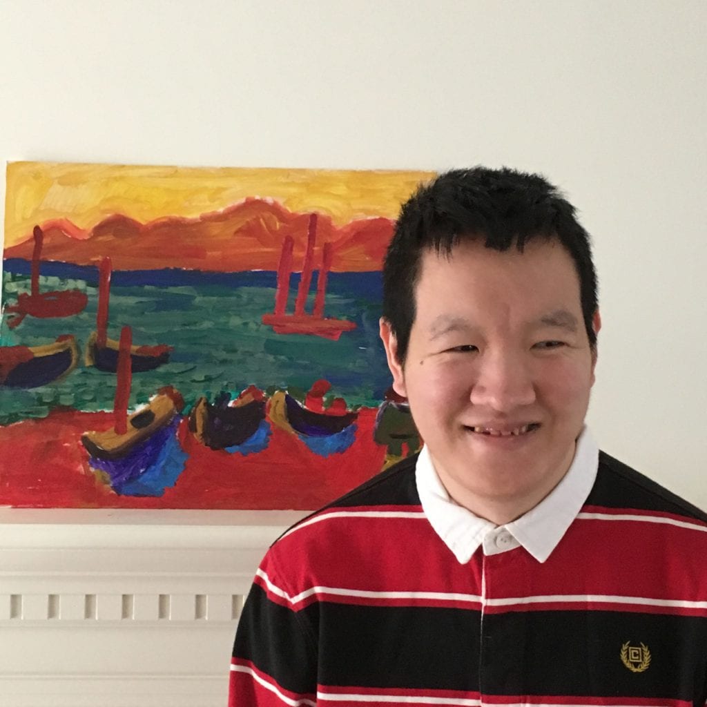 Headshot of a smiling man in a black and red striped polo with a paining of boats in the background.