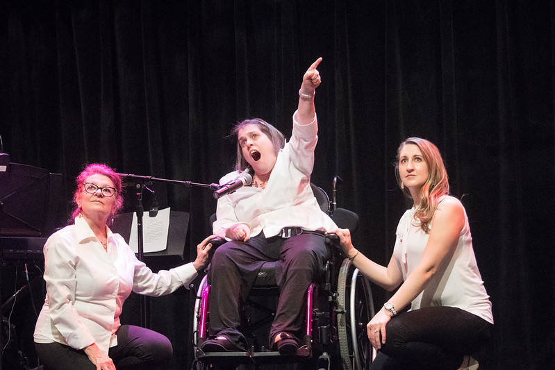 Three women wearing white tops and black pants on a stage. The woman in the middle is in a wheelchair and she is singing into a microphone while pointing up.