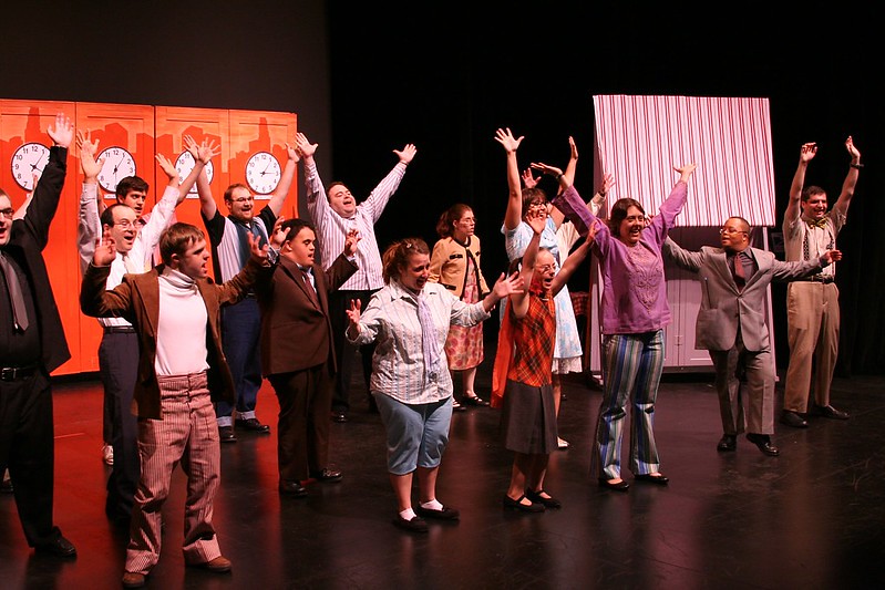 A group of actors on the stage with arms outstretched. The background has a set with clocks.