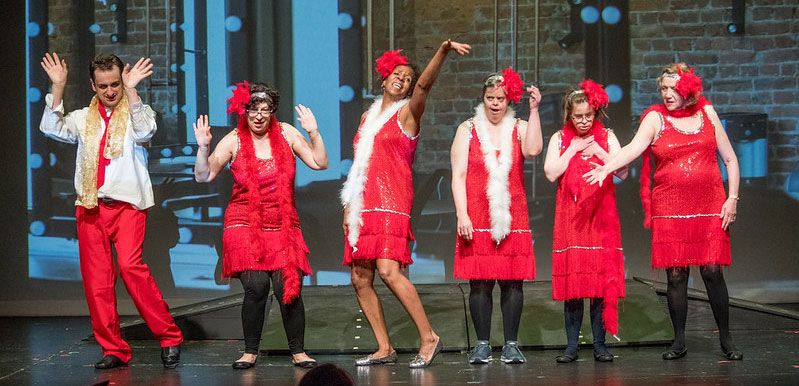 Group of actors on stage wearing red outfits and white boas